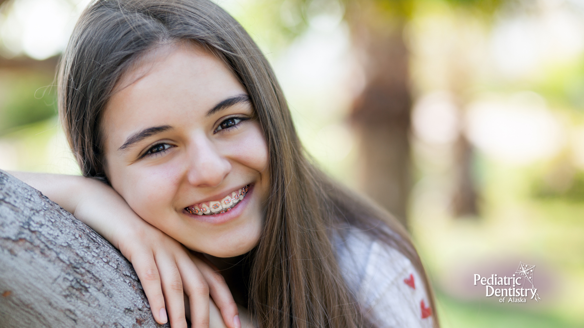 A young girl with braces is leaning on a tree trunk and smiling.