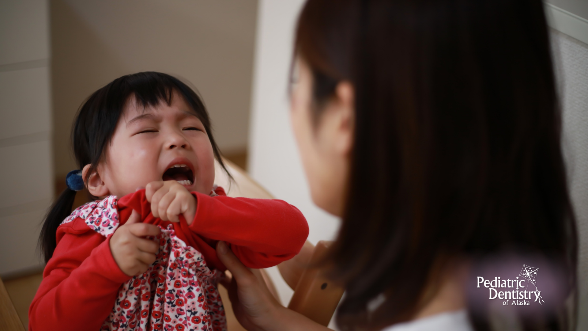 Crying child pulling at shirt, facing away from adult. Indoor setting, red and white clothing.