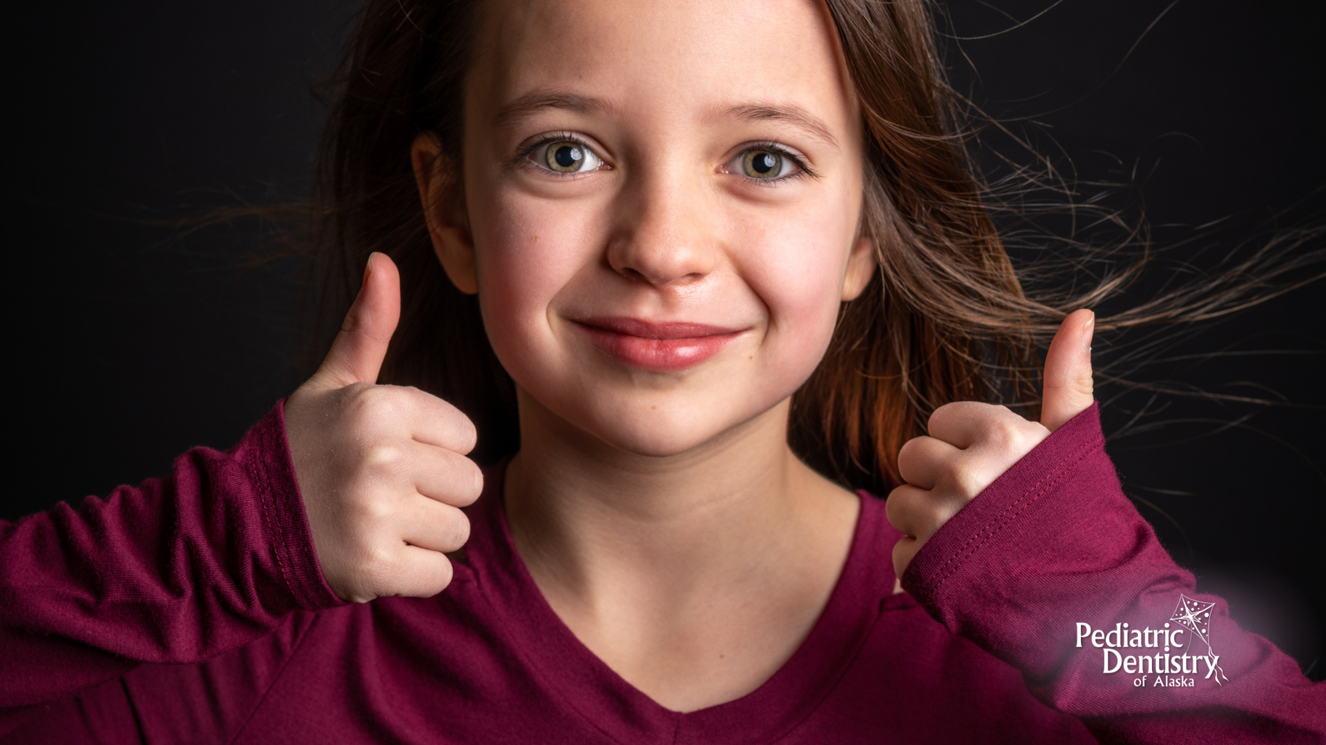 Girl with light skin, thumbs up, smiling, maroon shirt, black background.