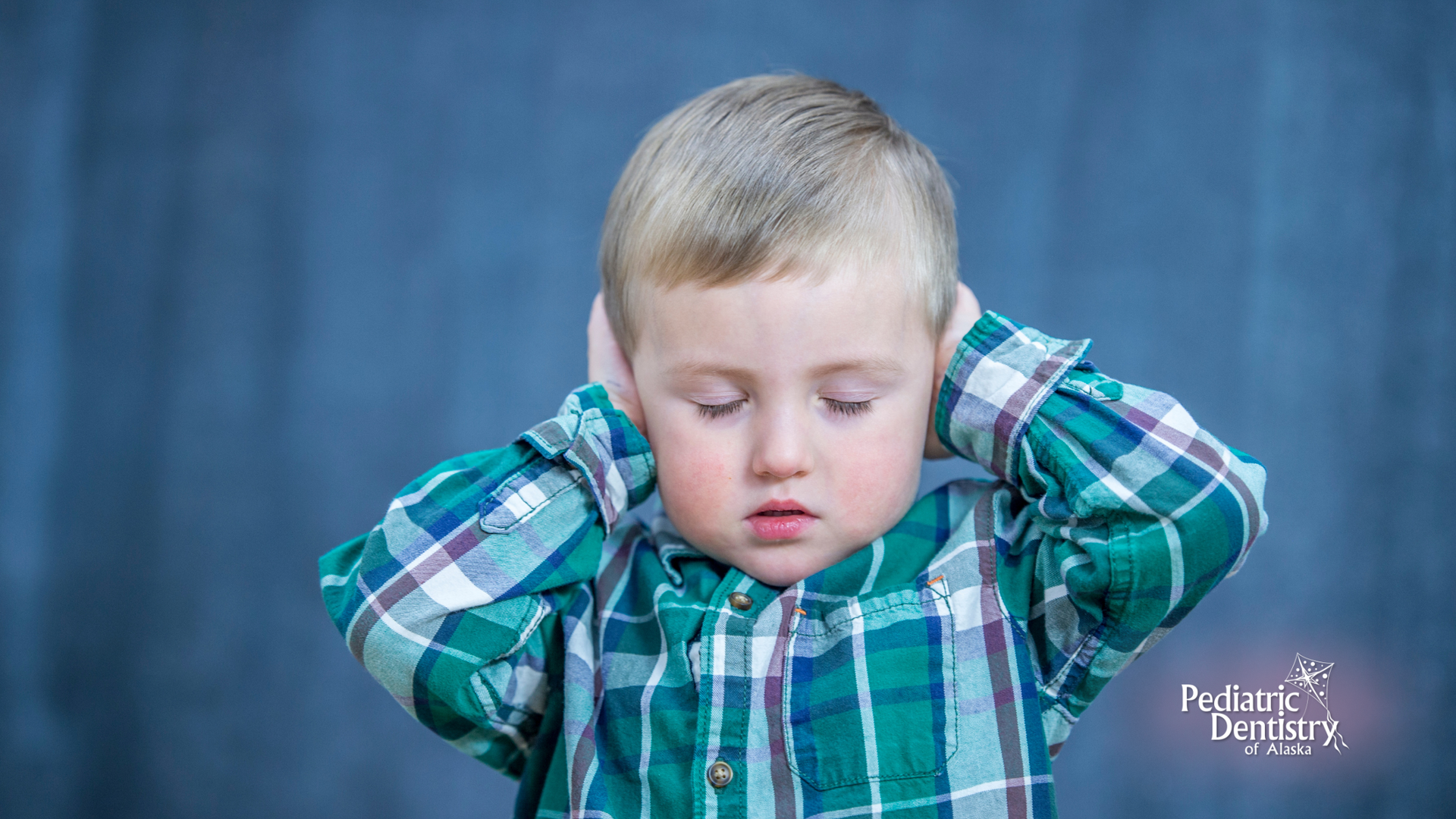 A young boy in a plaid shirt is covering his ears with his hands.