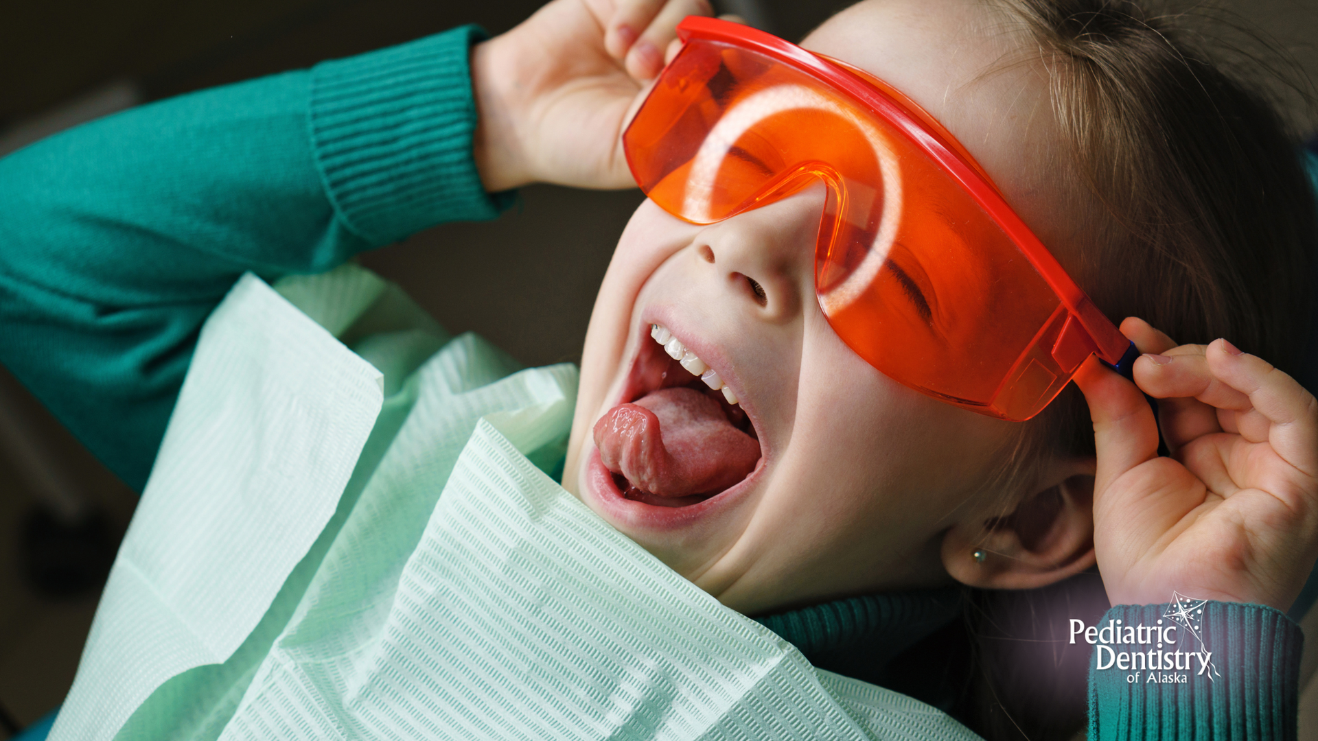 Child in dentist chair with mouth open, wearing orange safety glasses.
