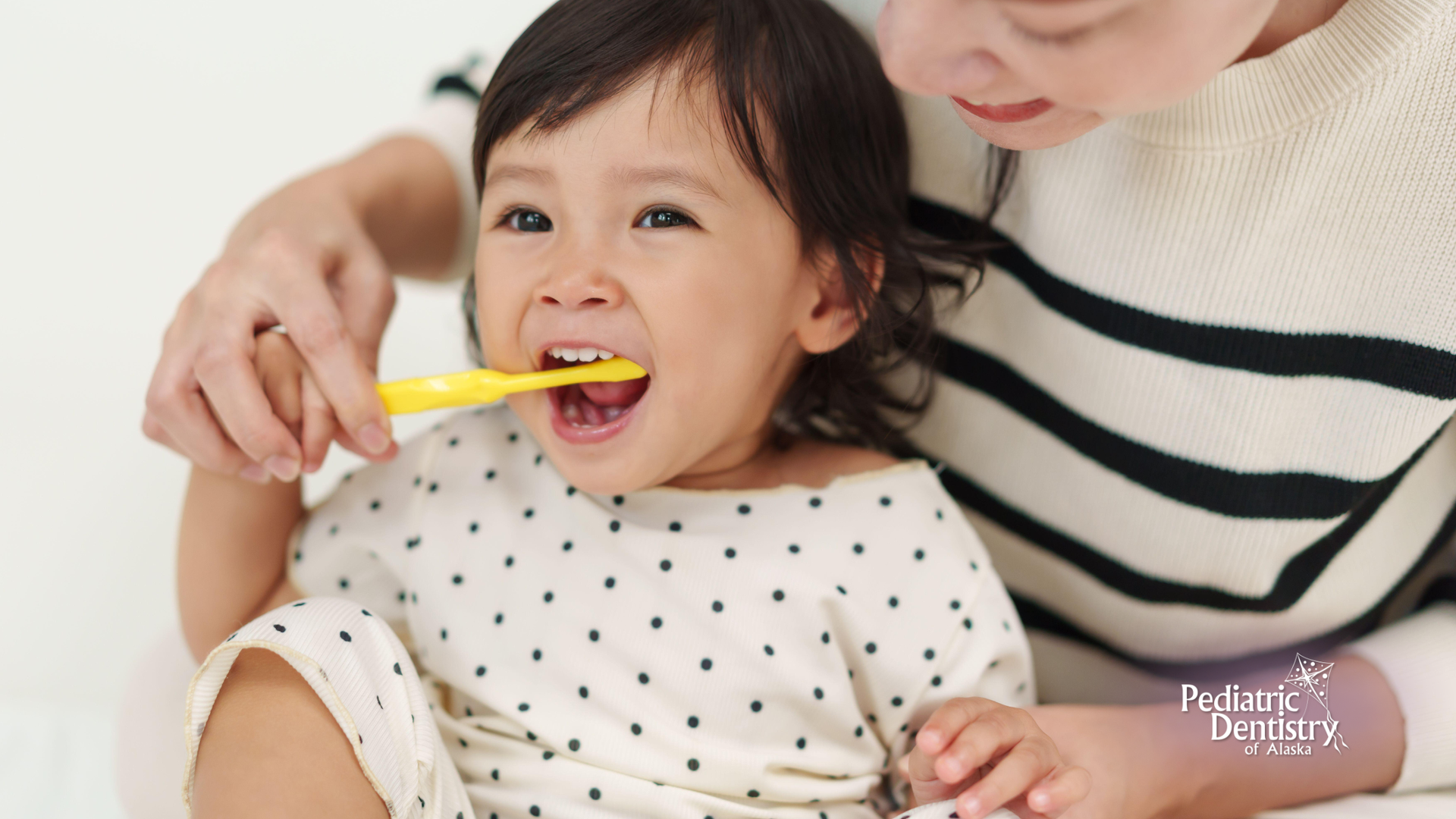 A person brushing a child's teeth with a yellow toothbrush; both are smiling.
