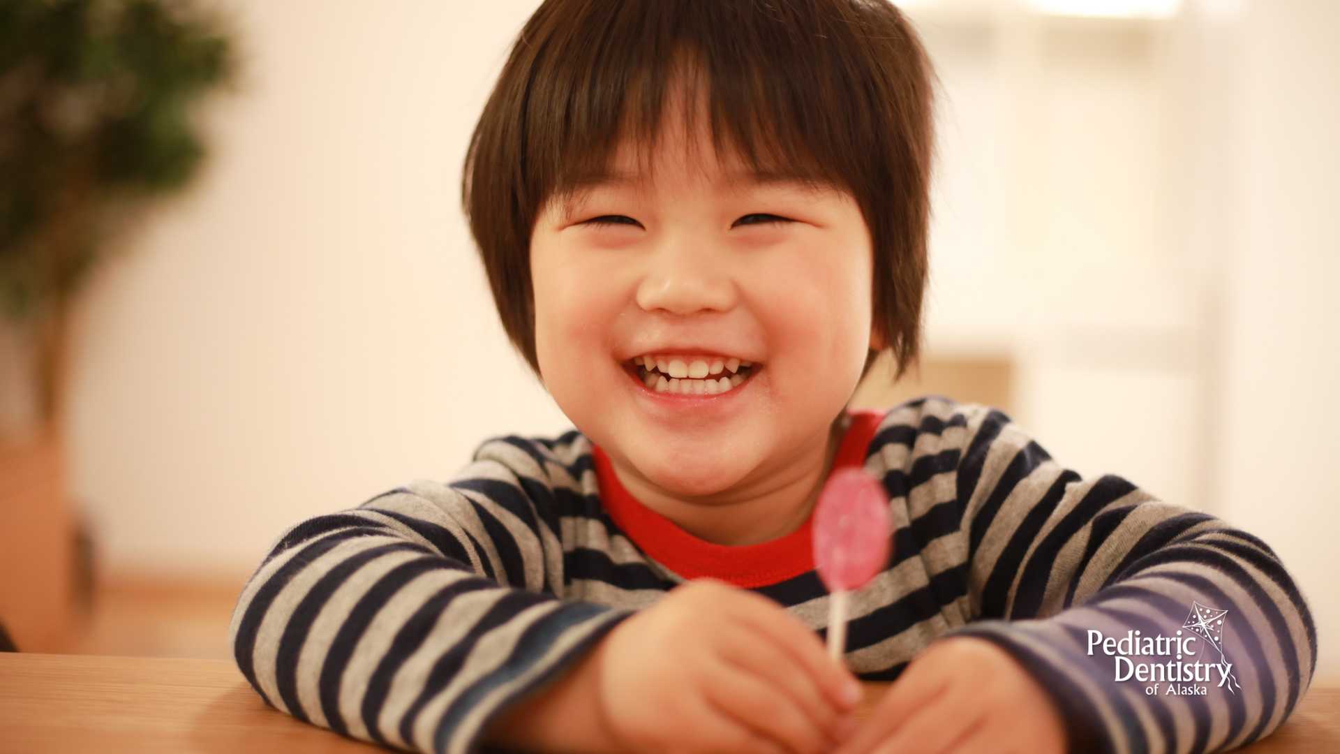 Smiling child holding a pink lollipop. Wearing striped shirt, sitting indoors, soft lighting.