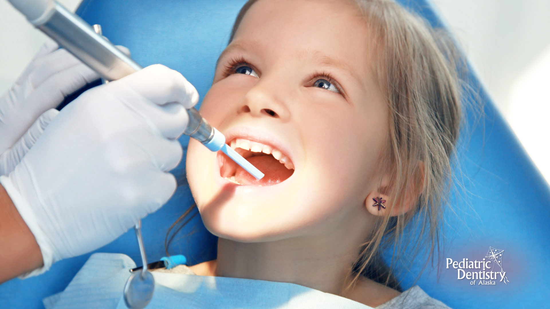 Child receiving dental care; dentist's hands and tools visible, blue setting.