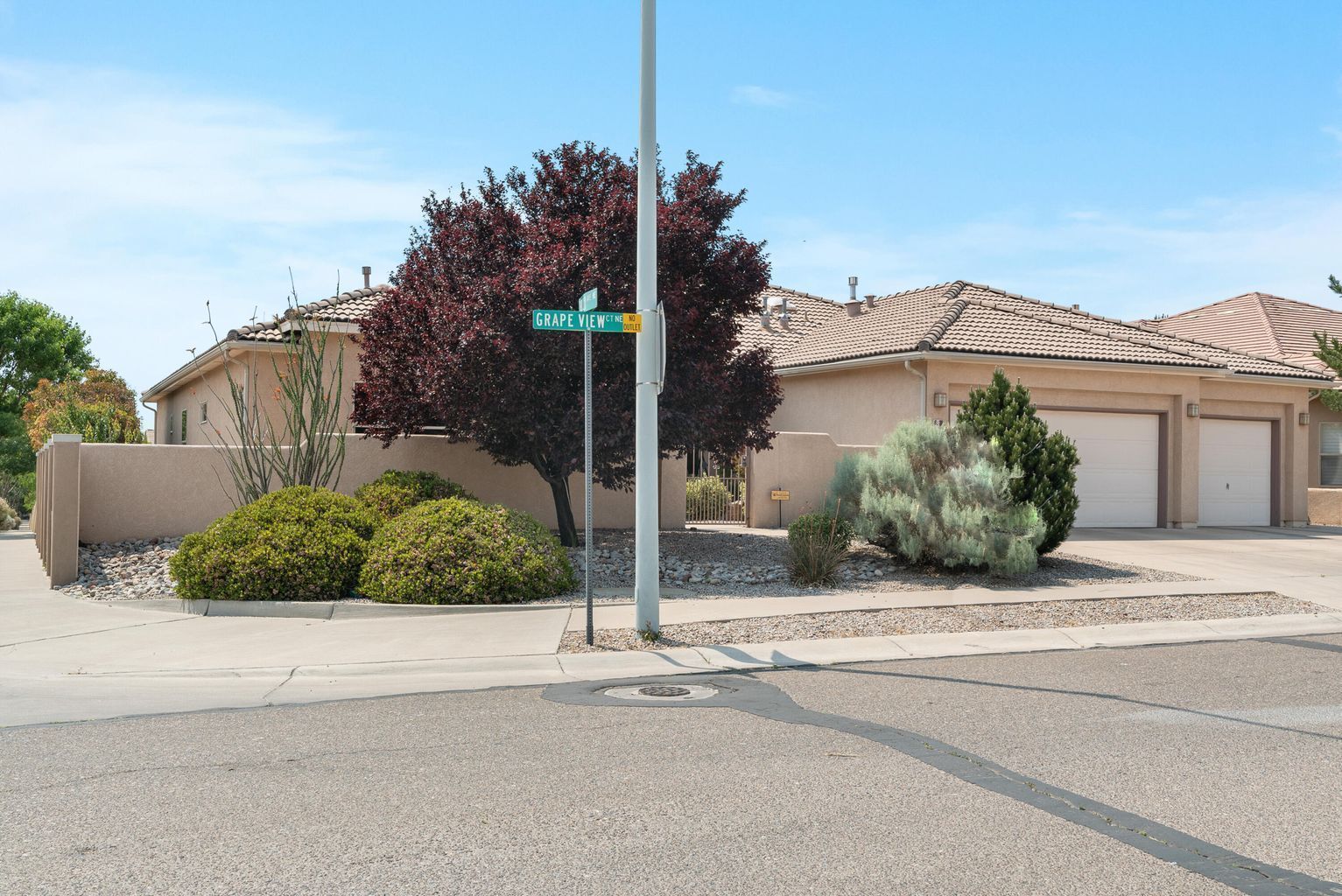 Beige stucco home with a two-car garage, red tree, street sign, and various bushes under a blue sky.