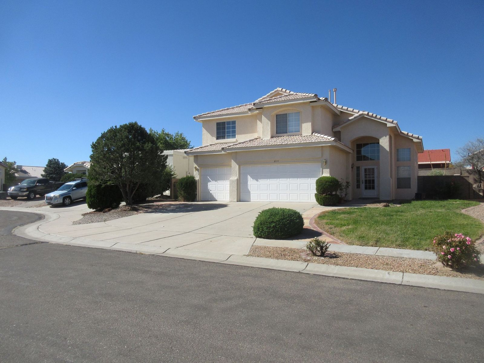 Two-story beige house with a two-car garage, on a paved driveway, in a suburban neighborhood on a sunny day.