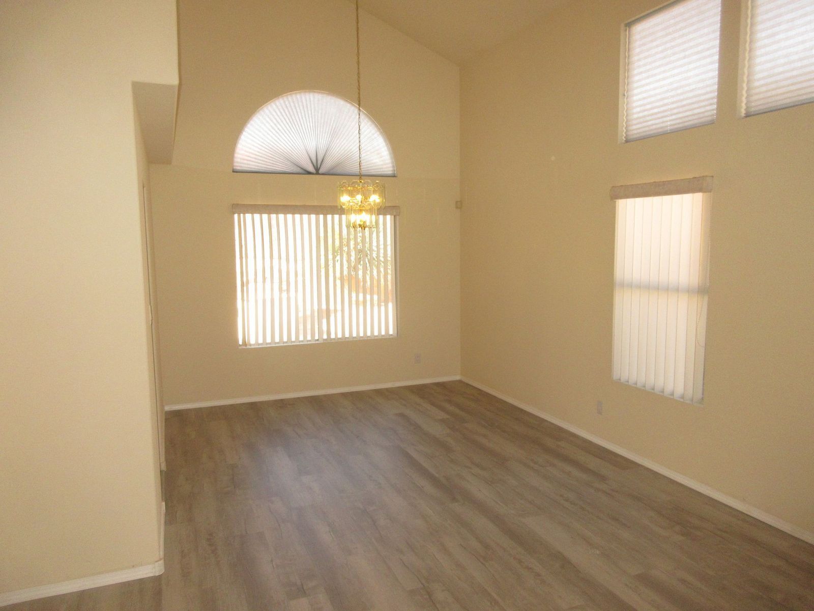 Empty living room with tall ceilings, windows with blinds, and wood-look flooring.