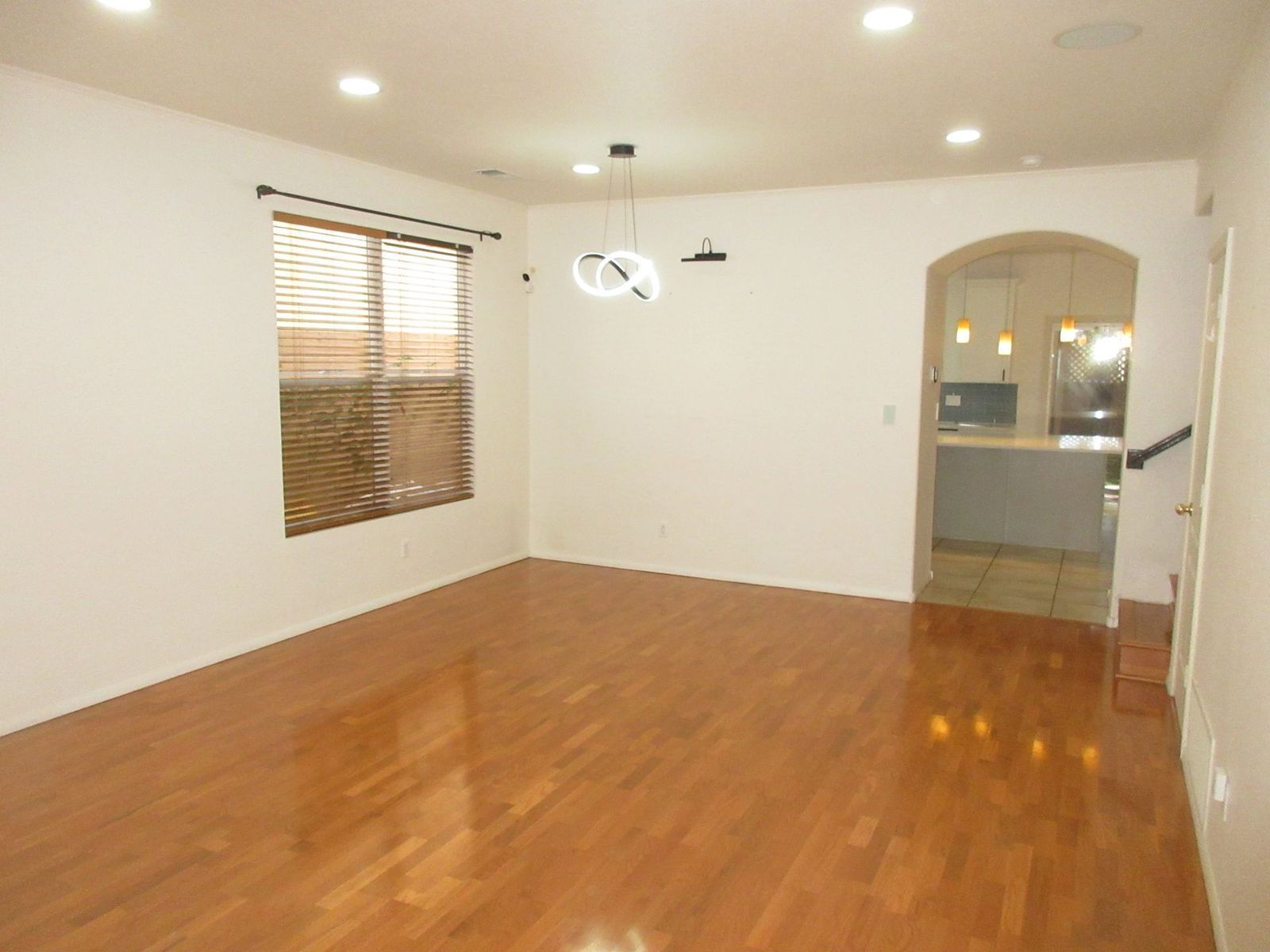 Empty dining room with hardwood floors, white walls, window with blinds, and arched doorway to a kitchen.