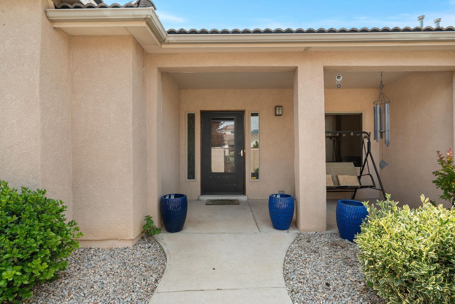 Entryway with a beige stucco exterior, dark door, blue planters, and a small sitting area with chimes.