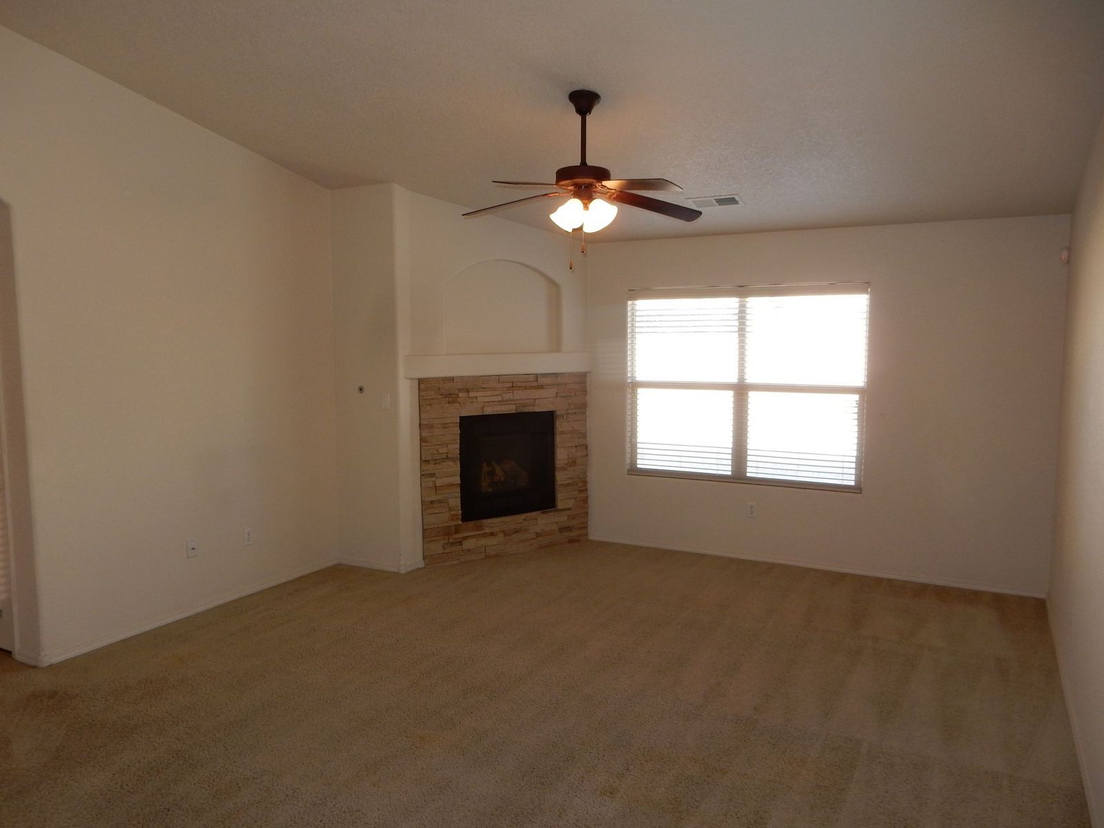 Empty living room with tan carpet, a stone fireplace, and a ceiling fan. A window provides natural light.