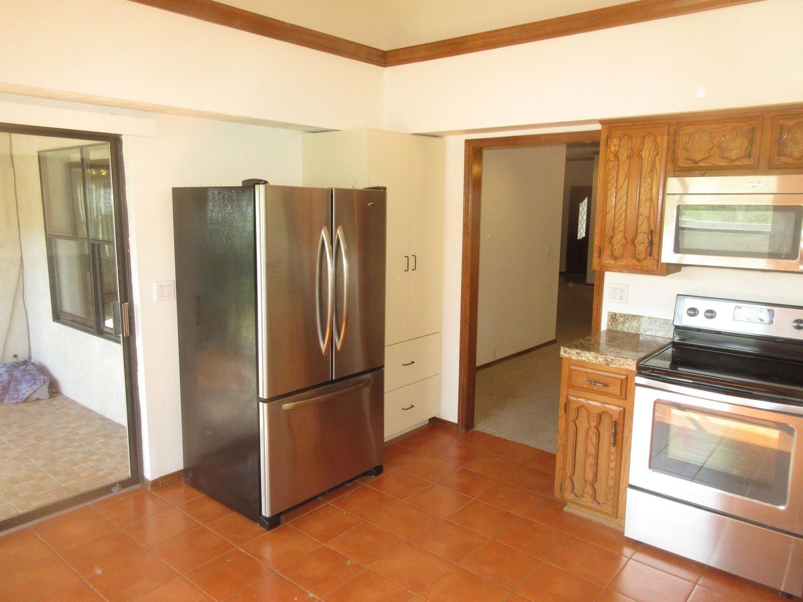 Kitchen with stainless steel refrigerator, brown cabinets, stove, and sliding door. Brown tiled floor.