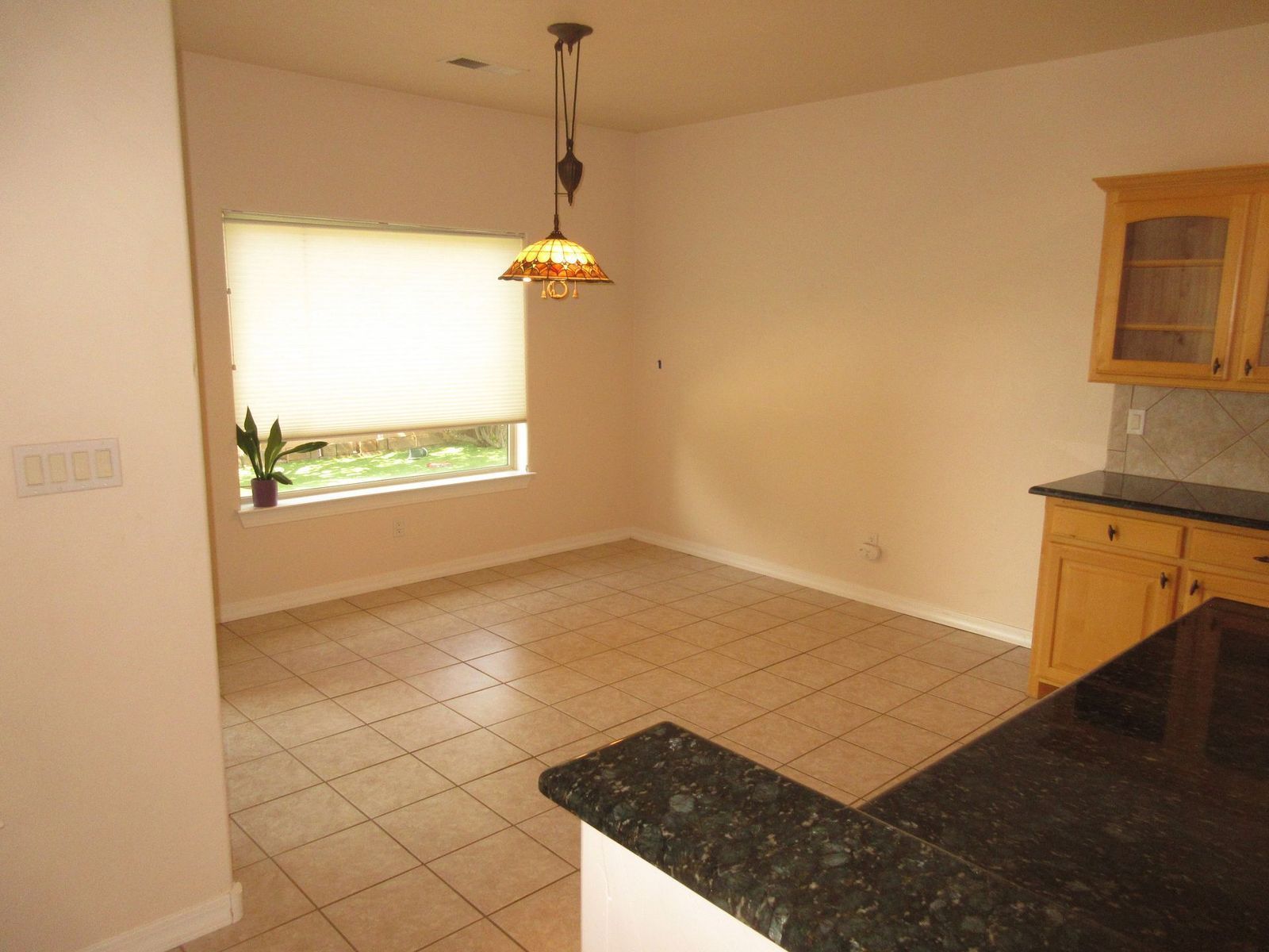Empty dining area with tiled floor, window, and overhead light fixture. Kitchen cabinets visible.