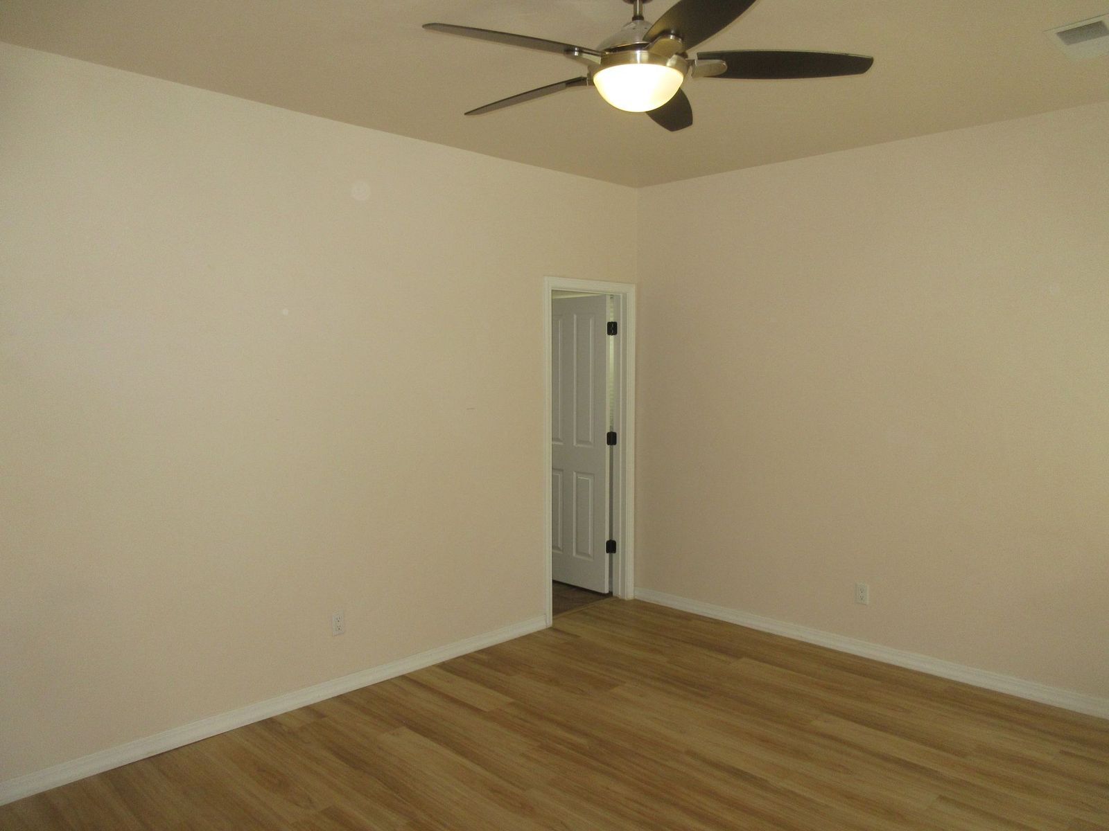 Empty room with tan walls, wood-look floor, and ceiling fan; a door is in the distance.