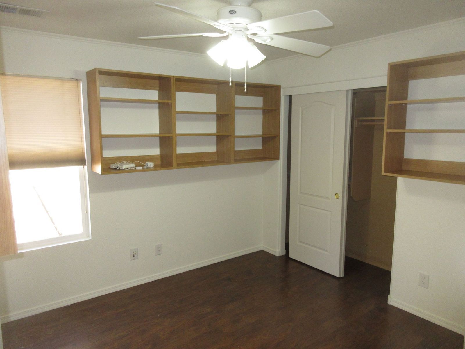 Empty bedroom with hardwood floor, built-in shelving, closet, and a ceiling fan.