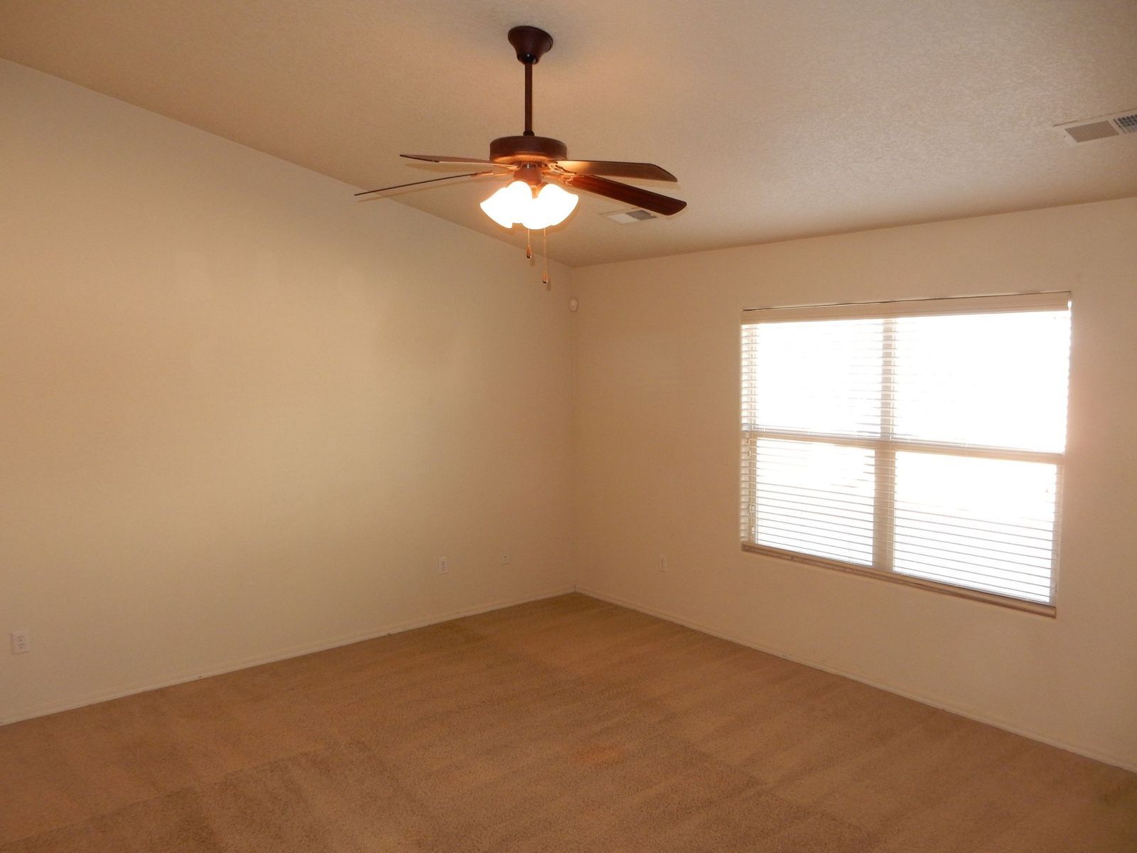 Empty room with tan carpet, beige walls, and a window with blinds; a ceiling fan is mounted.