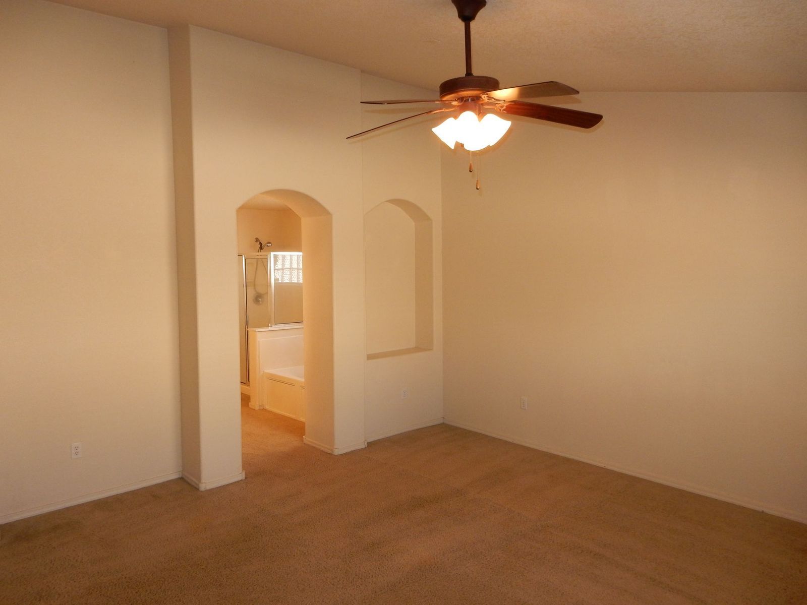 Empty bedroom with carpet, ceiling fan, and arched doorway leading to a bathroom. Beige walls and carpet.