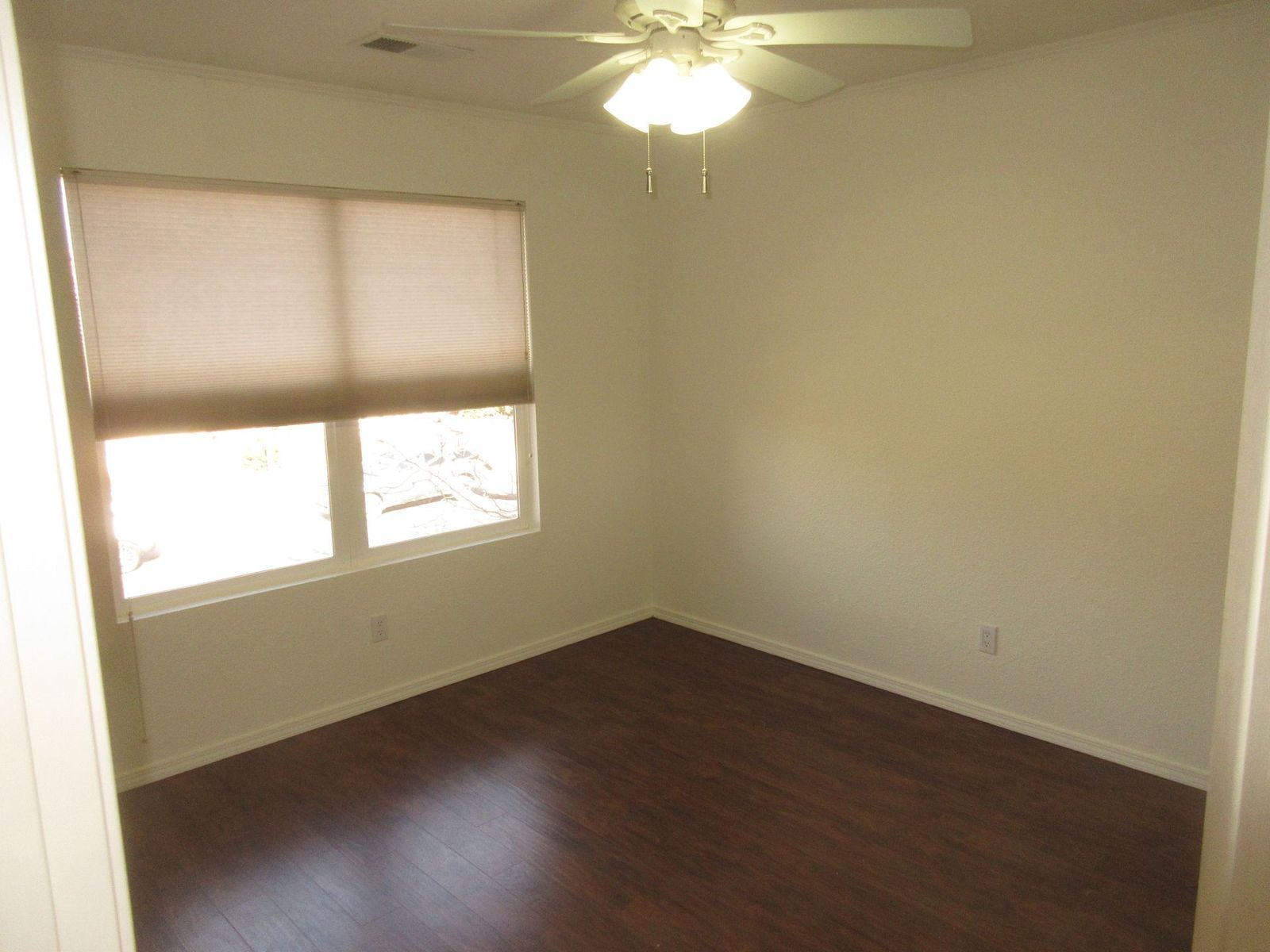 Empty room with dark wood flooring, window with blind, ceiling fan.