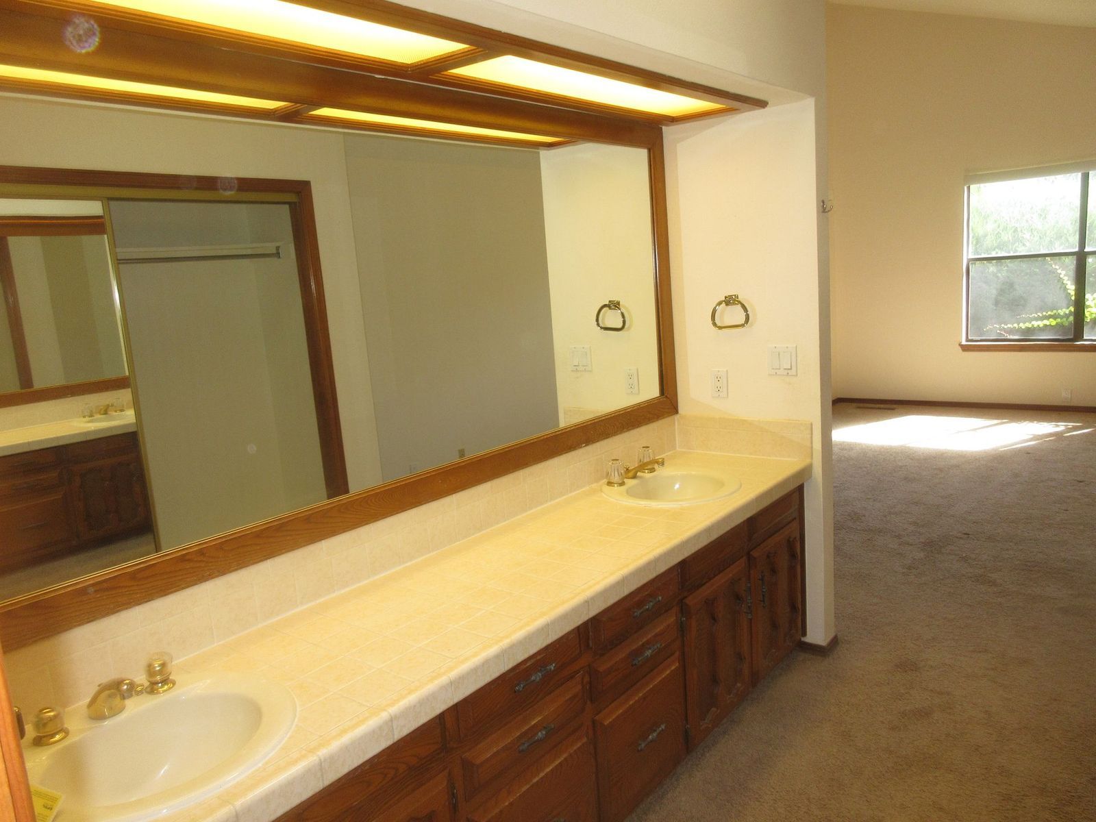 Bathroom with double vanity, large mirror, and open doorway to bedroom with window.