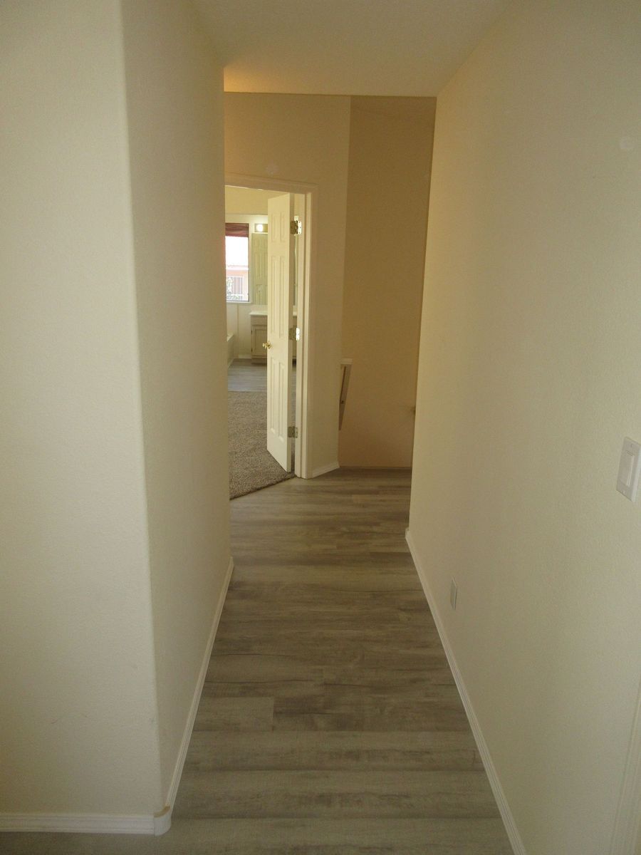 Narrow hallway with wood-look flooring and off-white walls. Doorway at the end leads to a room.