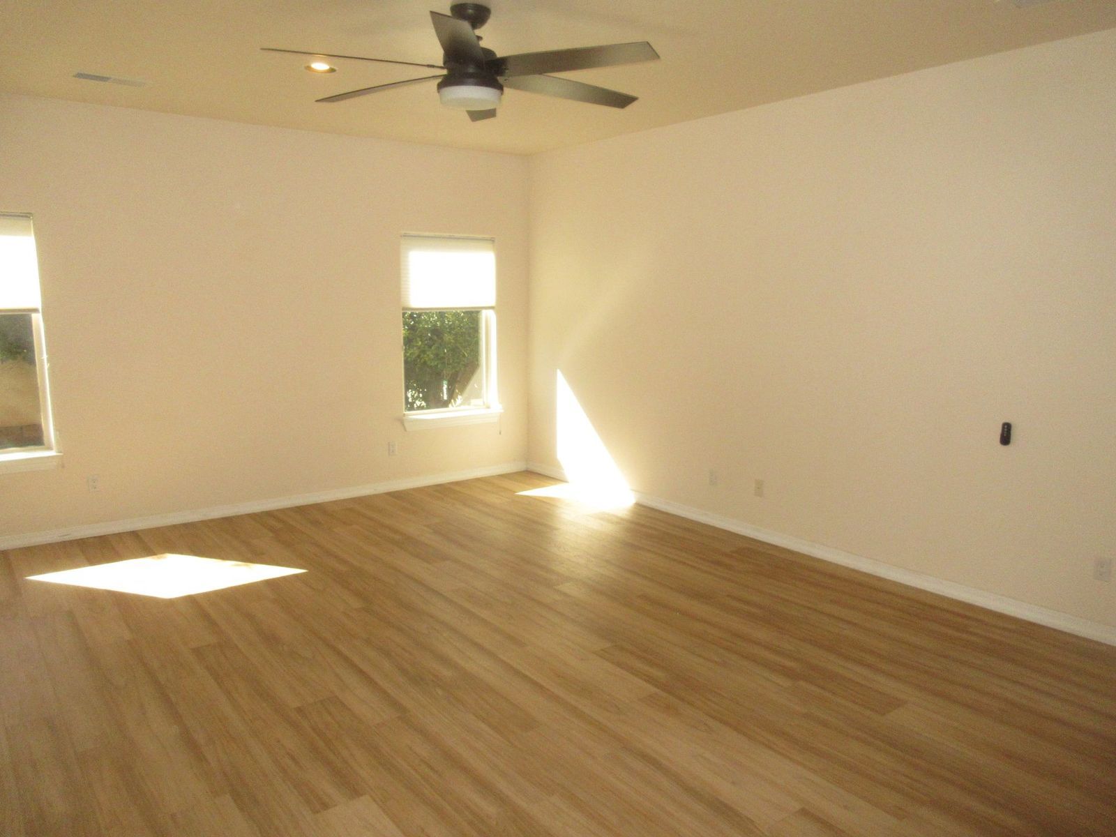 Empty room with hardwood floors, white walls, and a ceiling fan; sunlight streams in.