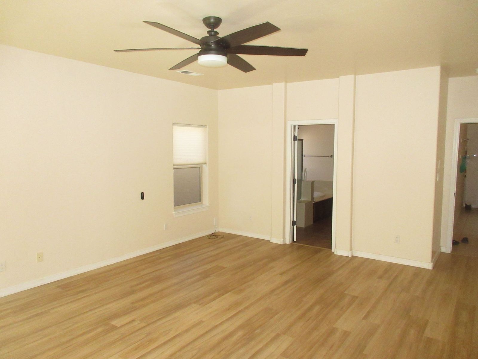 Empty room with light wood-look flooring, cream walls, and a ceiling fan. Doorway to bathroom visible.