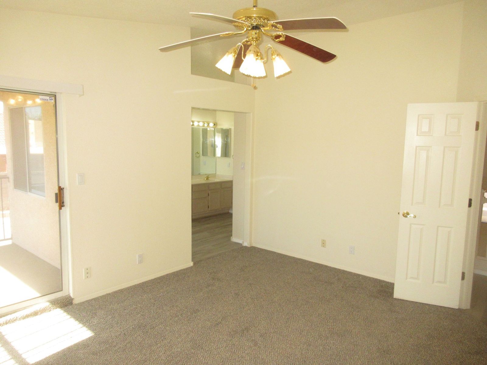 Empty beige-walled room with carpet, ceiling fan, open door, and doorway to bathroom.