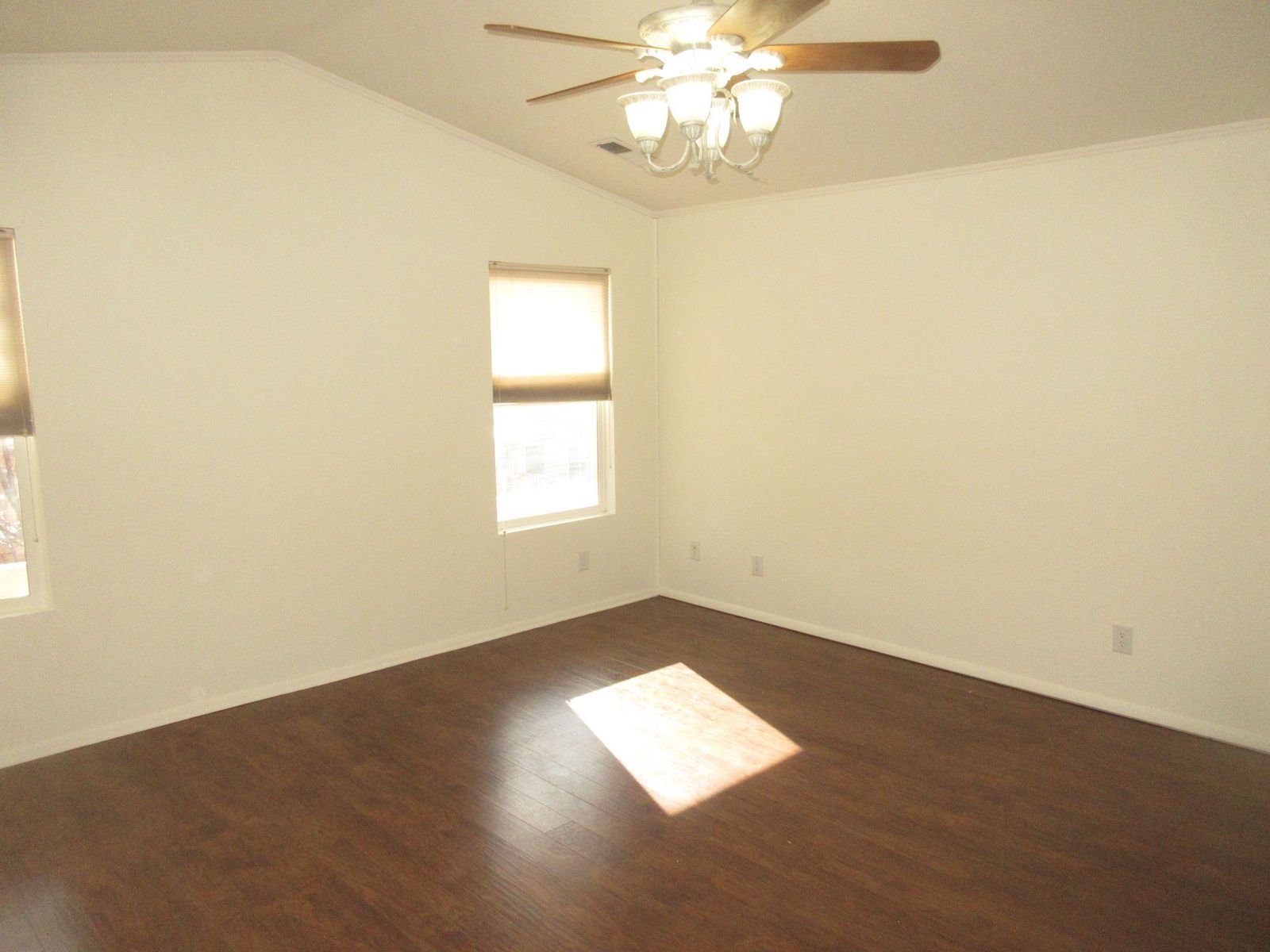 Empty room with dark wood flooring, angled ceiling, ceiling fan, and a window letting in sunlight.