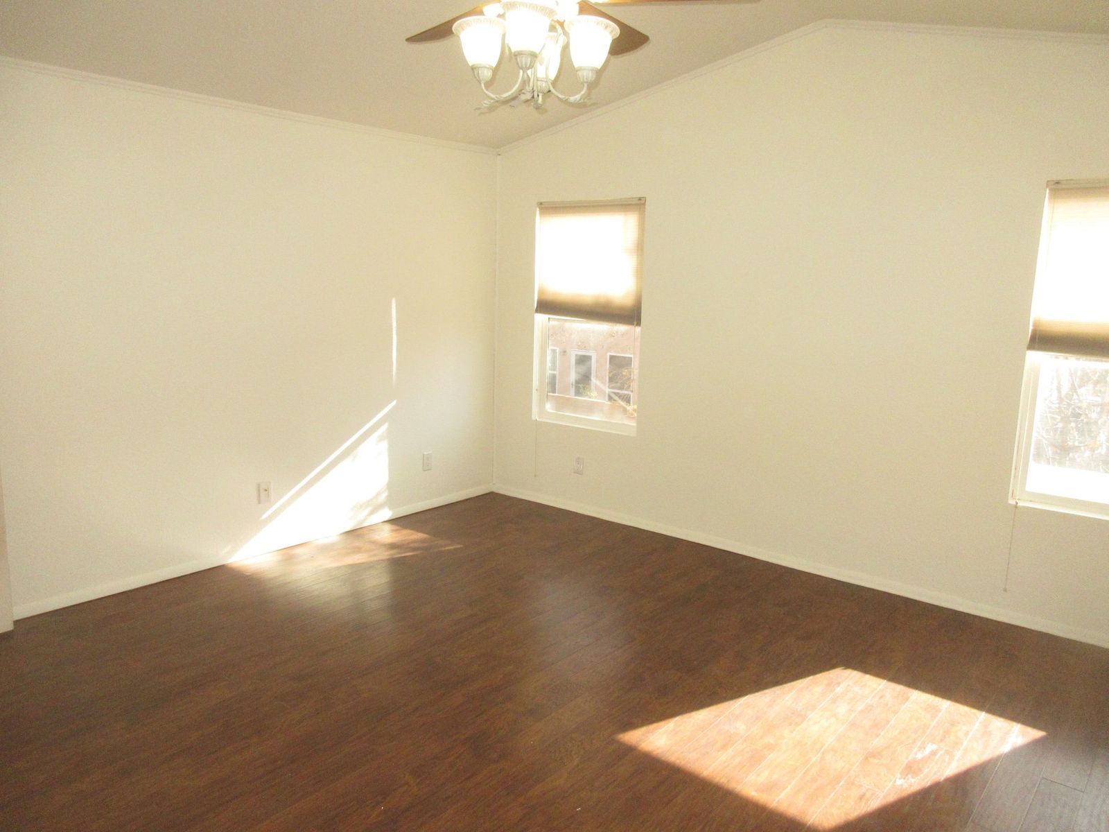 Empty room with dark wood floor, white walls, two windows with shades, ceiling fan, and sunlit square.