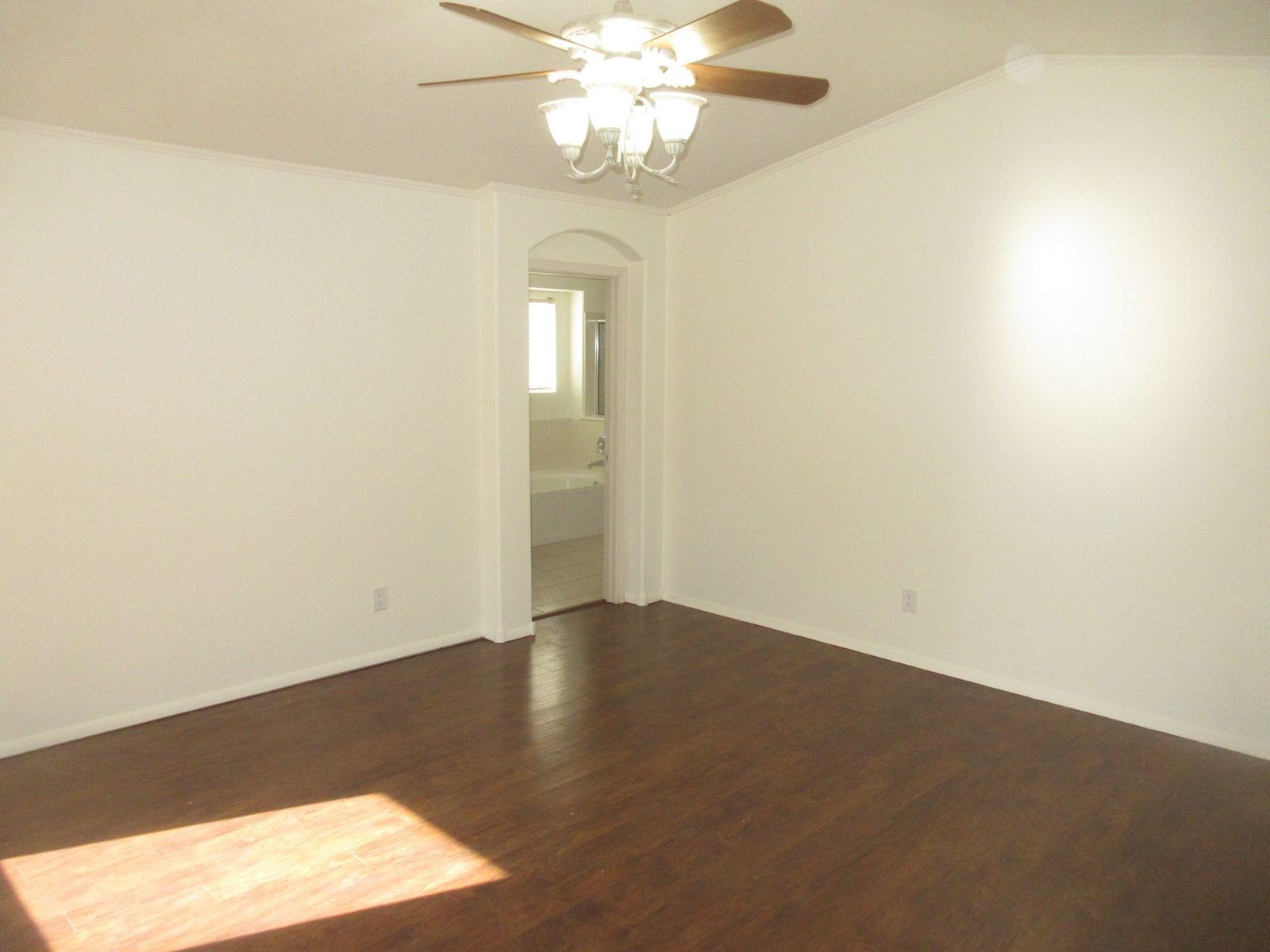 Empty room with brown hardwood floors, white walls, and a ceiling fan. An arched doorway leads to a bathroom.