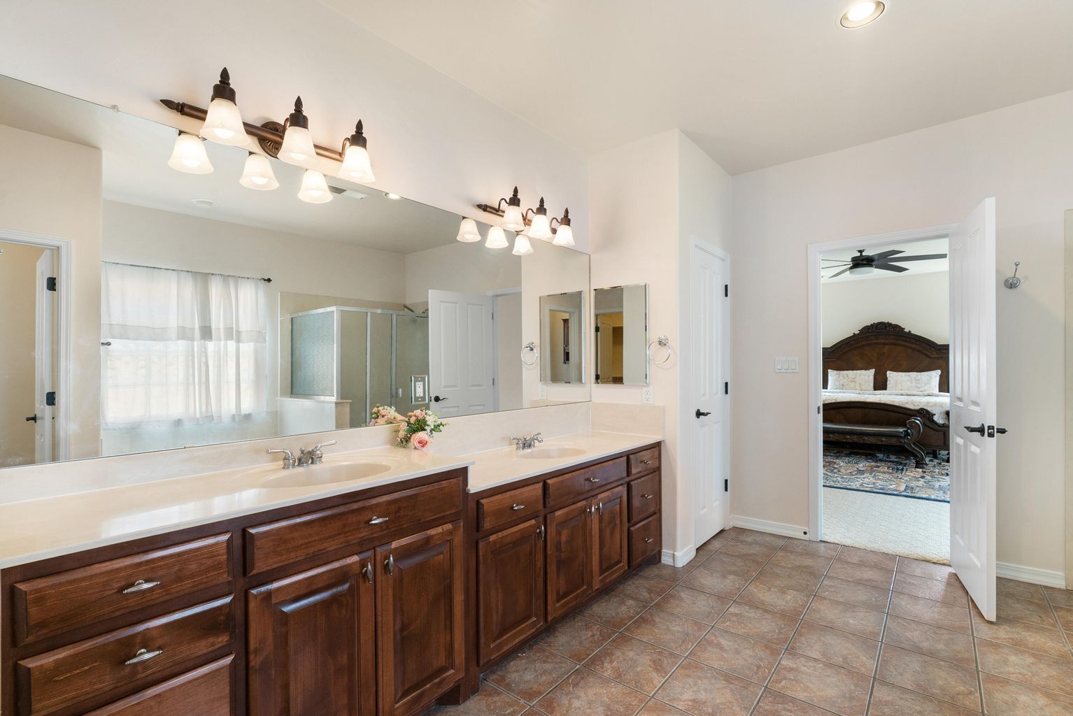 Bathroom with double vanity, dark cabinets, bright lighting, and an open doorway to a bedroom.