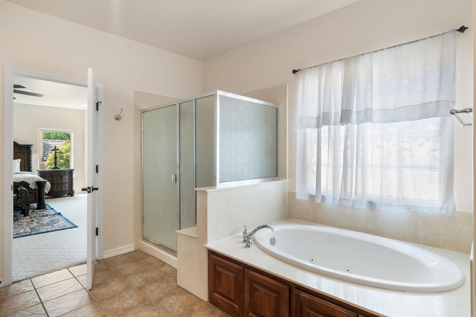 Bathroom with a tub, shower, and doorway to a bedroom. White walls, wood cabinets, and beige floor tiles.