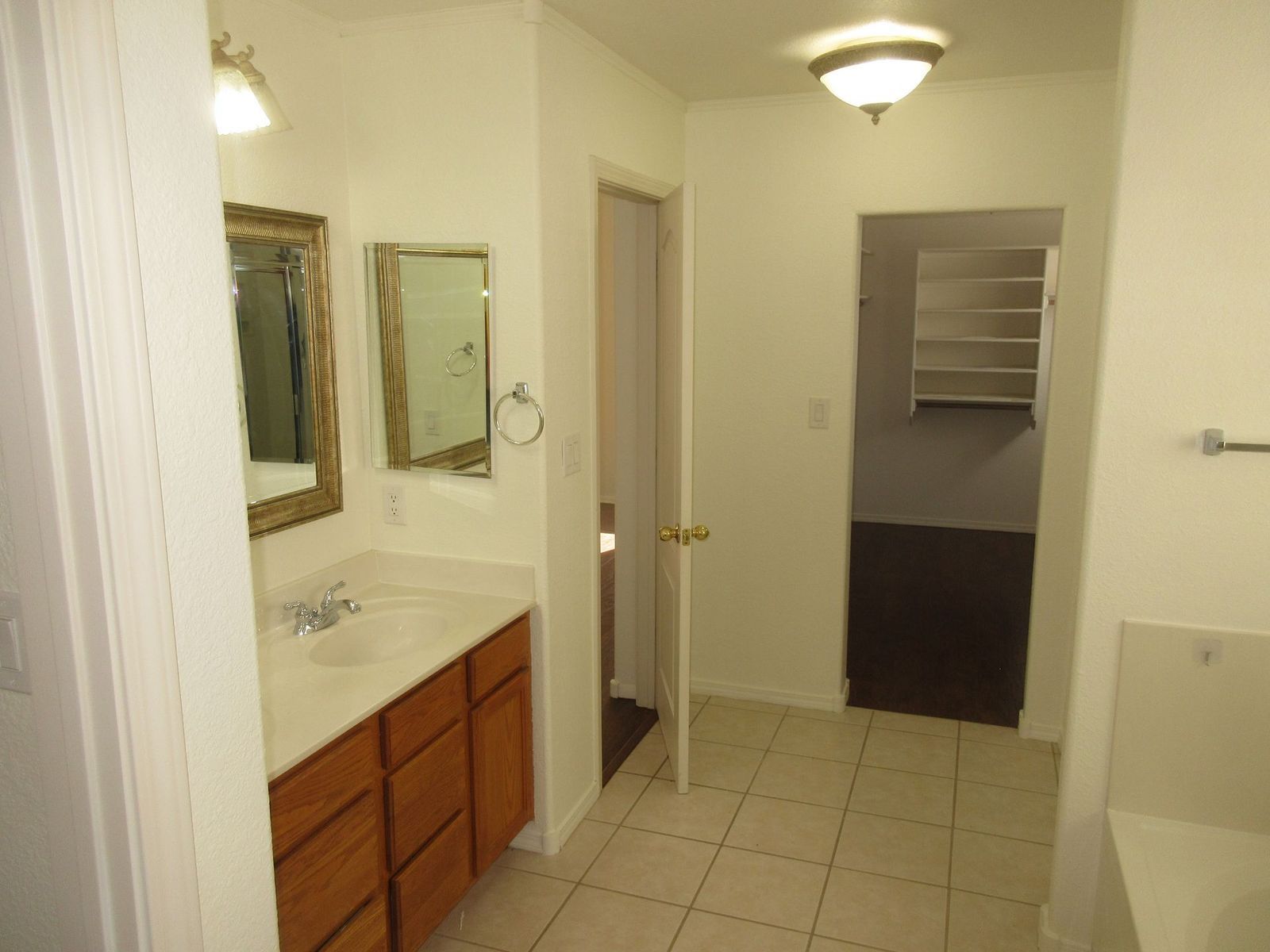 Bathroom with a vanity, mirror, and a hallway leading to a closet. Brown cabinets and beige tile.
