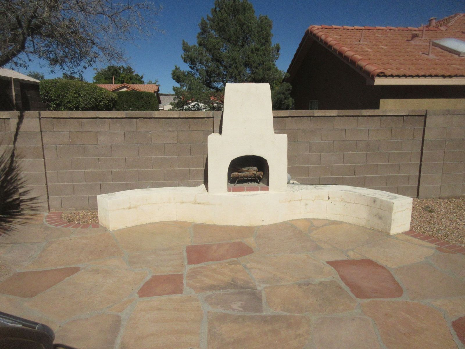 Outdoor fireplace with beige stucco surround against a cinder block wall, with a stone patio.