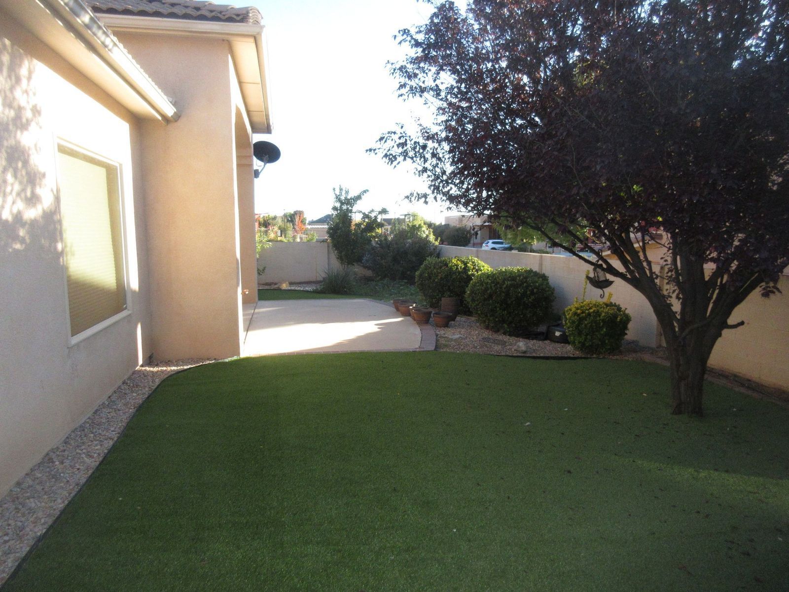 Lawn with artificial grass next to a house with a concrete patio and a tree in the corner, sunny day.