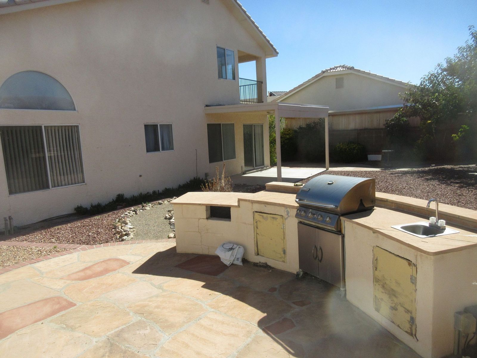 Backyard with outdoor kitchen and patio, next to a beige house, with blue sky.