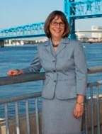 Woman in a blue suit smiles, standing near water with a bridge in the background.