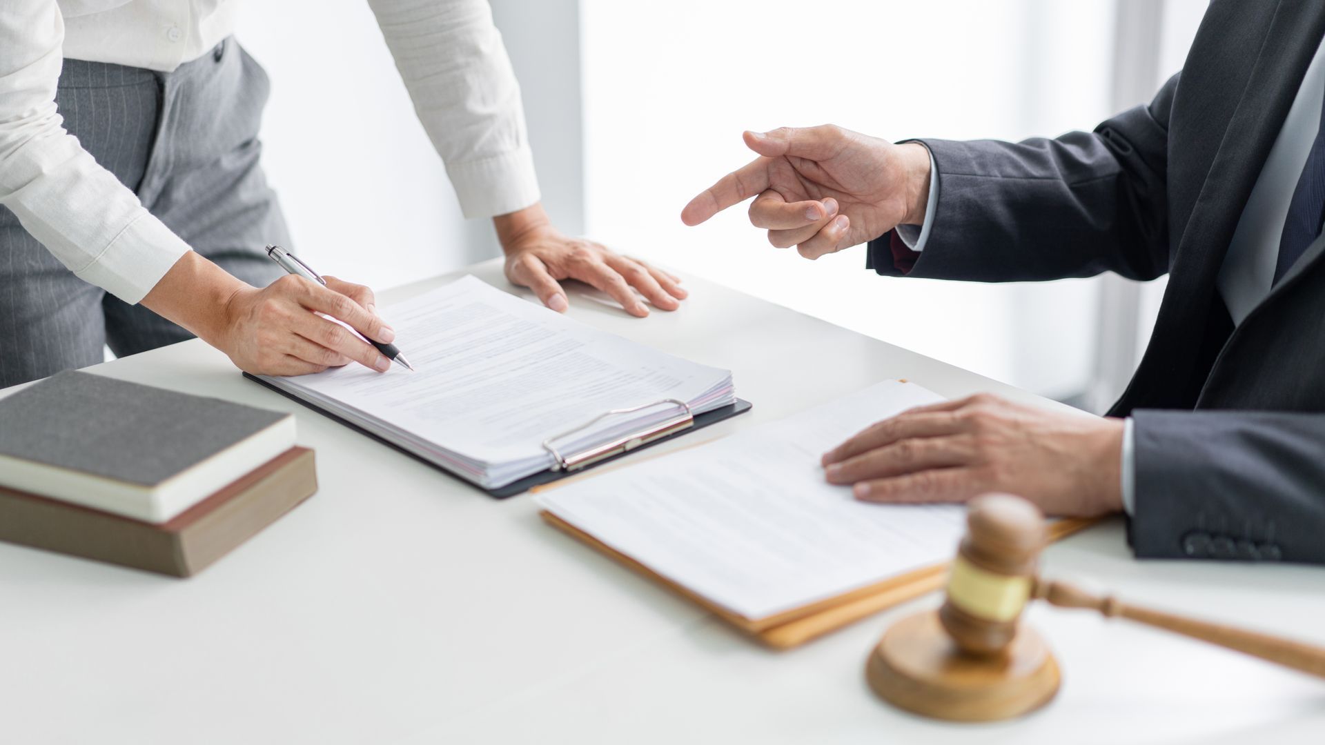 Person signing document with another pointing. Gavel and books on table.