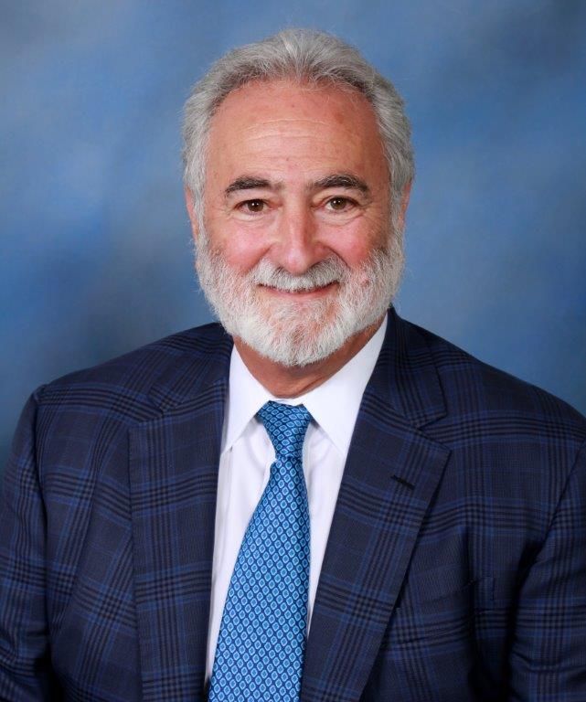 Man in a blue plaid suit and tie, smiling, with a grey beard, against a blue background.