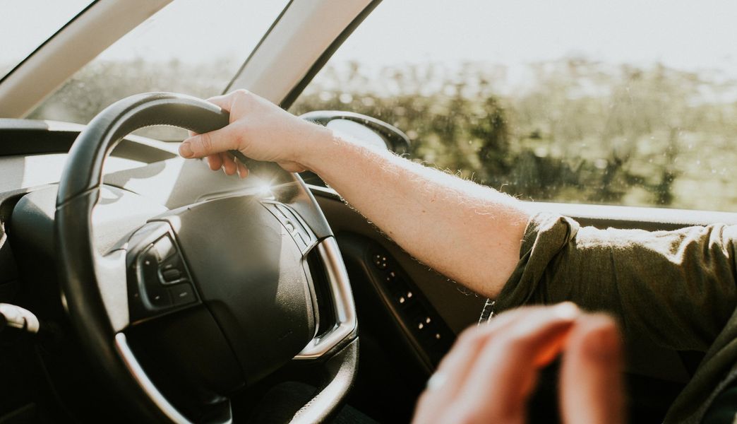 Person's hands gripping steering wheel, driving a car on a sunny day.