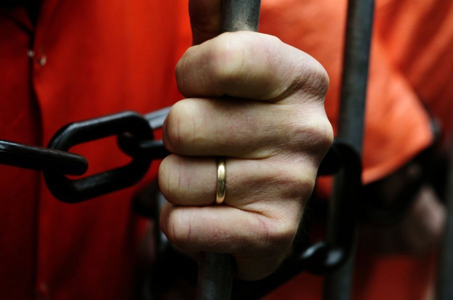 Close-up of a hand gripping prison bars; wearing an orange jumpsuit and a gold ring.
