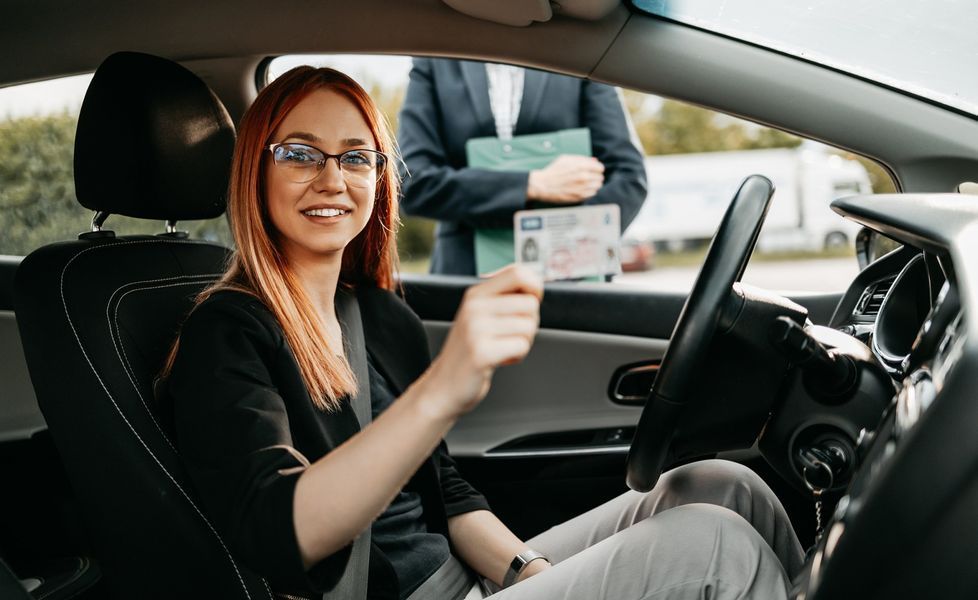 Woman in car holding up driver's license, smiling. Person in background holds clipboard.