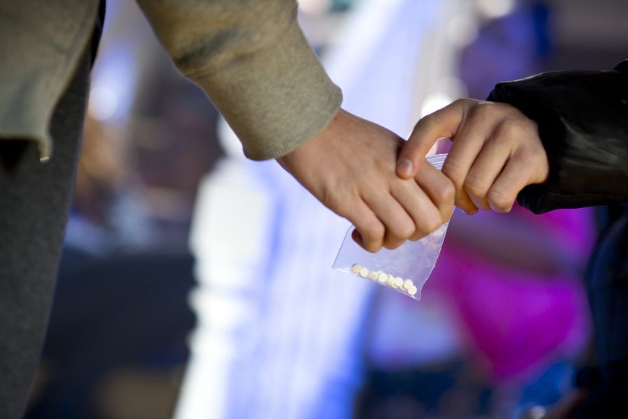 Two hands exchanging a small bag with white pills in a brightly lit setting, possibly a club.