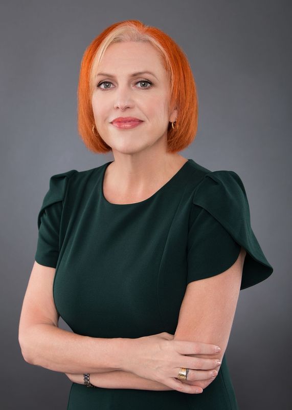 Woman with orange-red hair in green dress, arms crossed, smiling. Gray background.