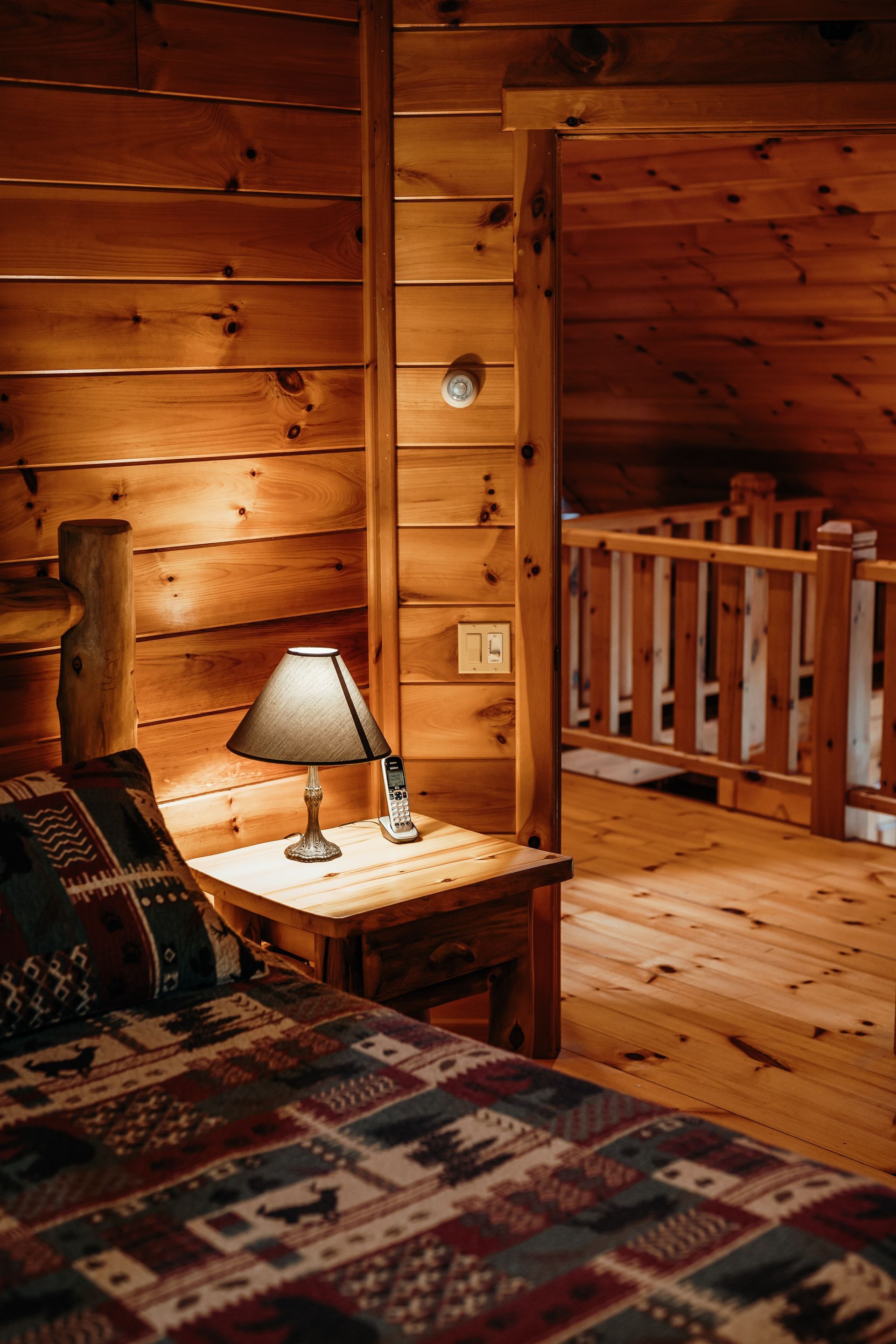 A bedroom in a log cabin with a bed , nightstand , lamp and stairs.