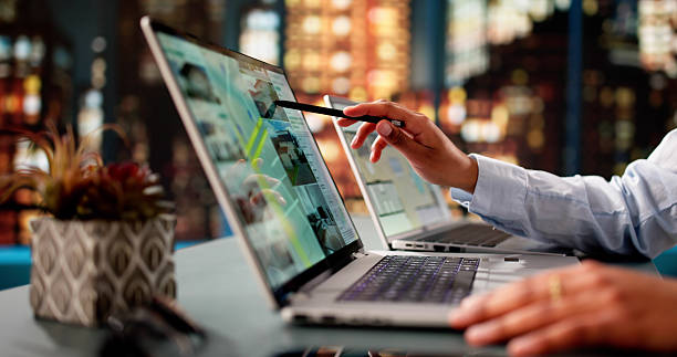 Realtor  points to a video call screen on a laptop. Other laptop visible. Soft lighting and city vie