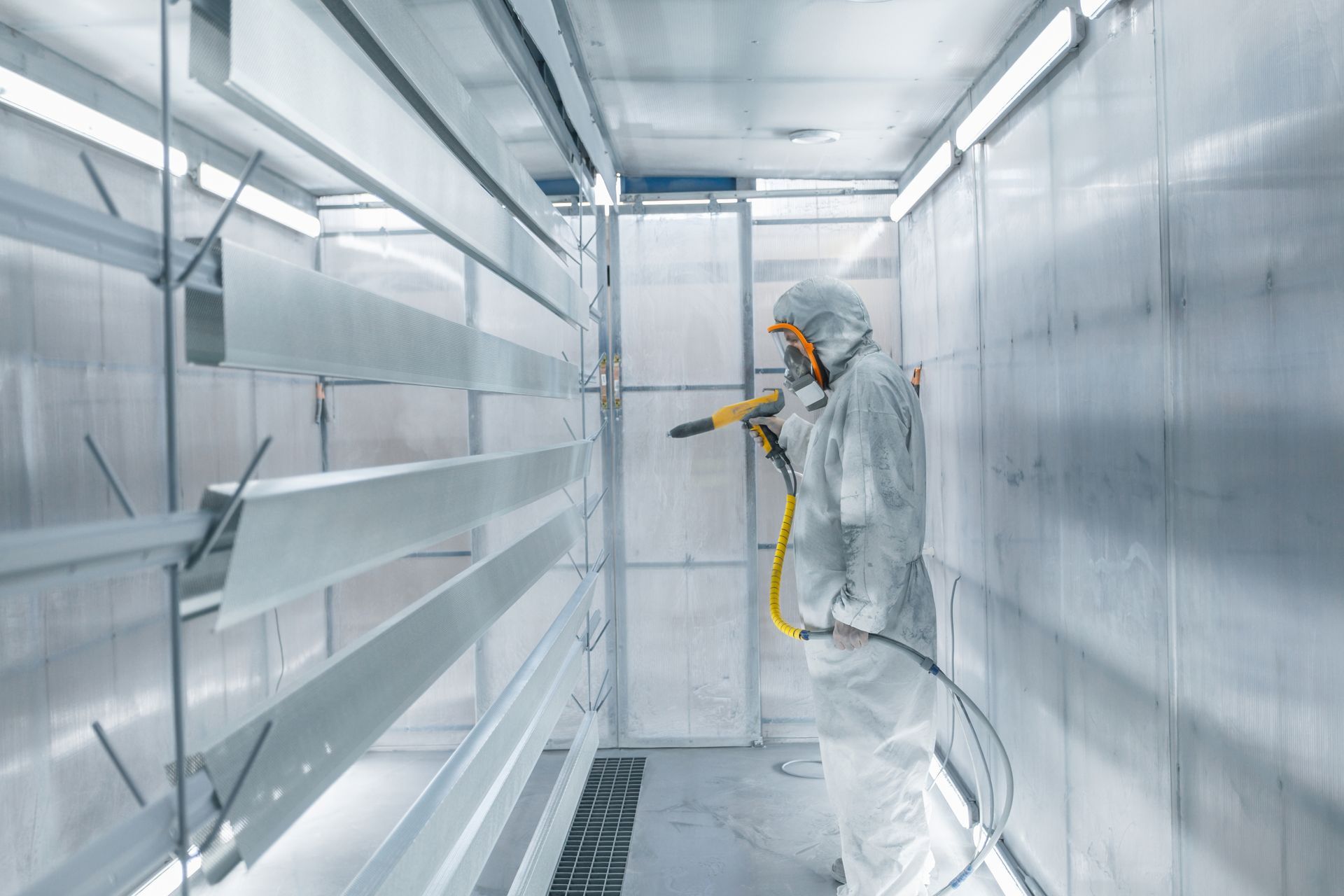 A person in a protective suit spray-paints metal parts inside a bright, industrial booth.