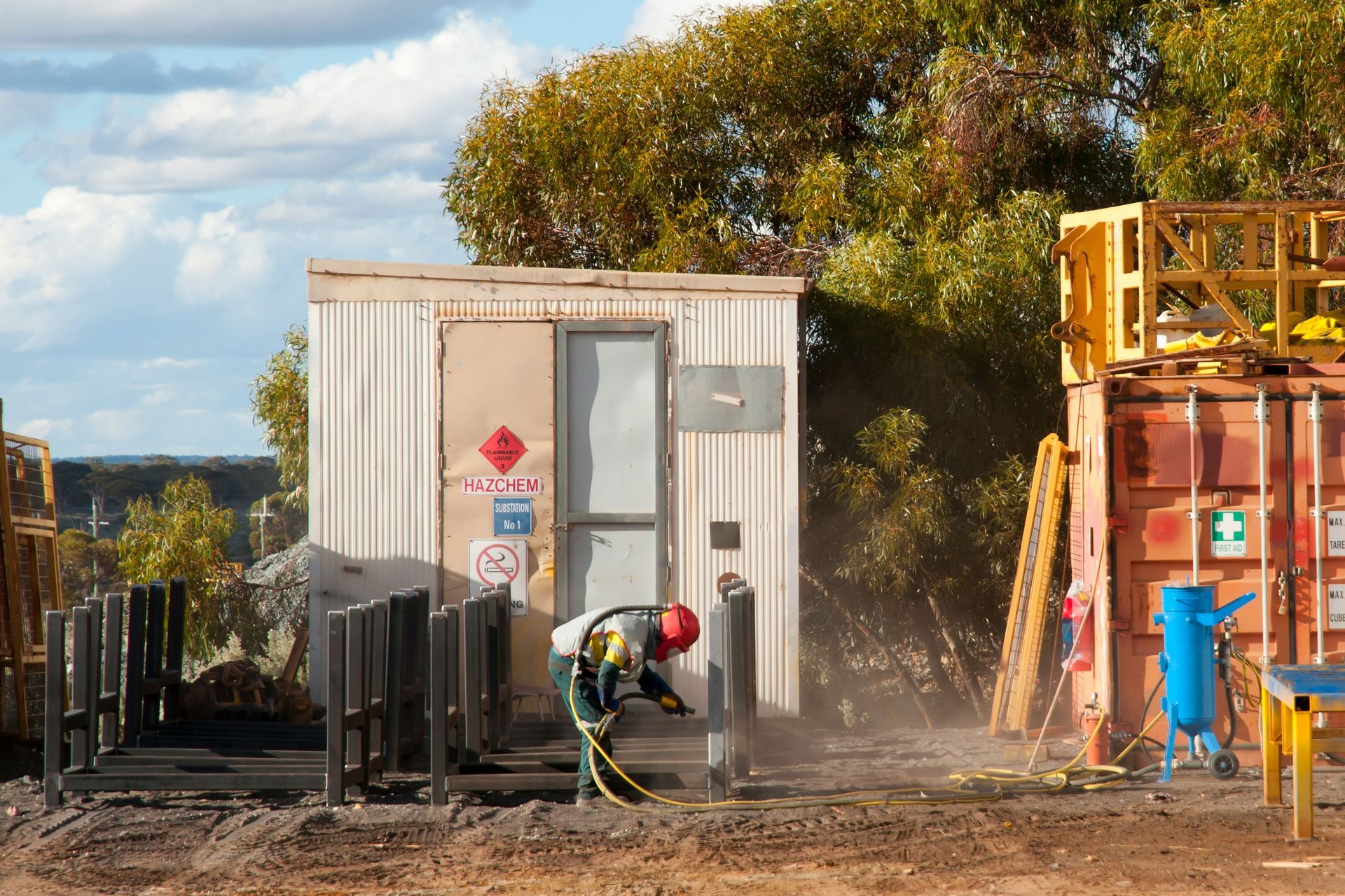 Worker in protective gear spraying a metal structure near a small building and containers in a rural setting.