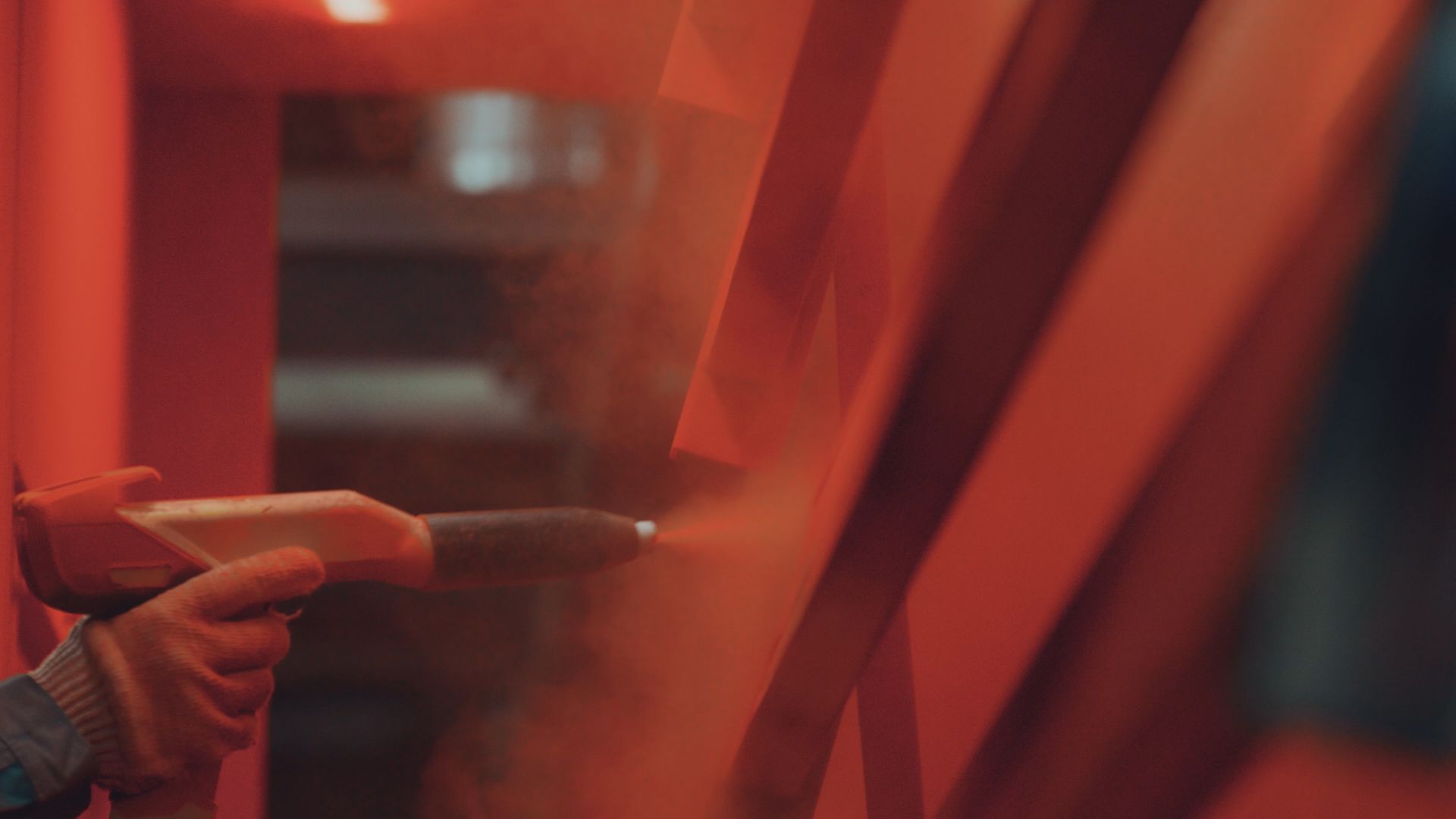 A person using a spray gun to apply orange powder coat to metal beams.