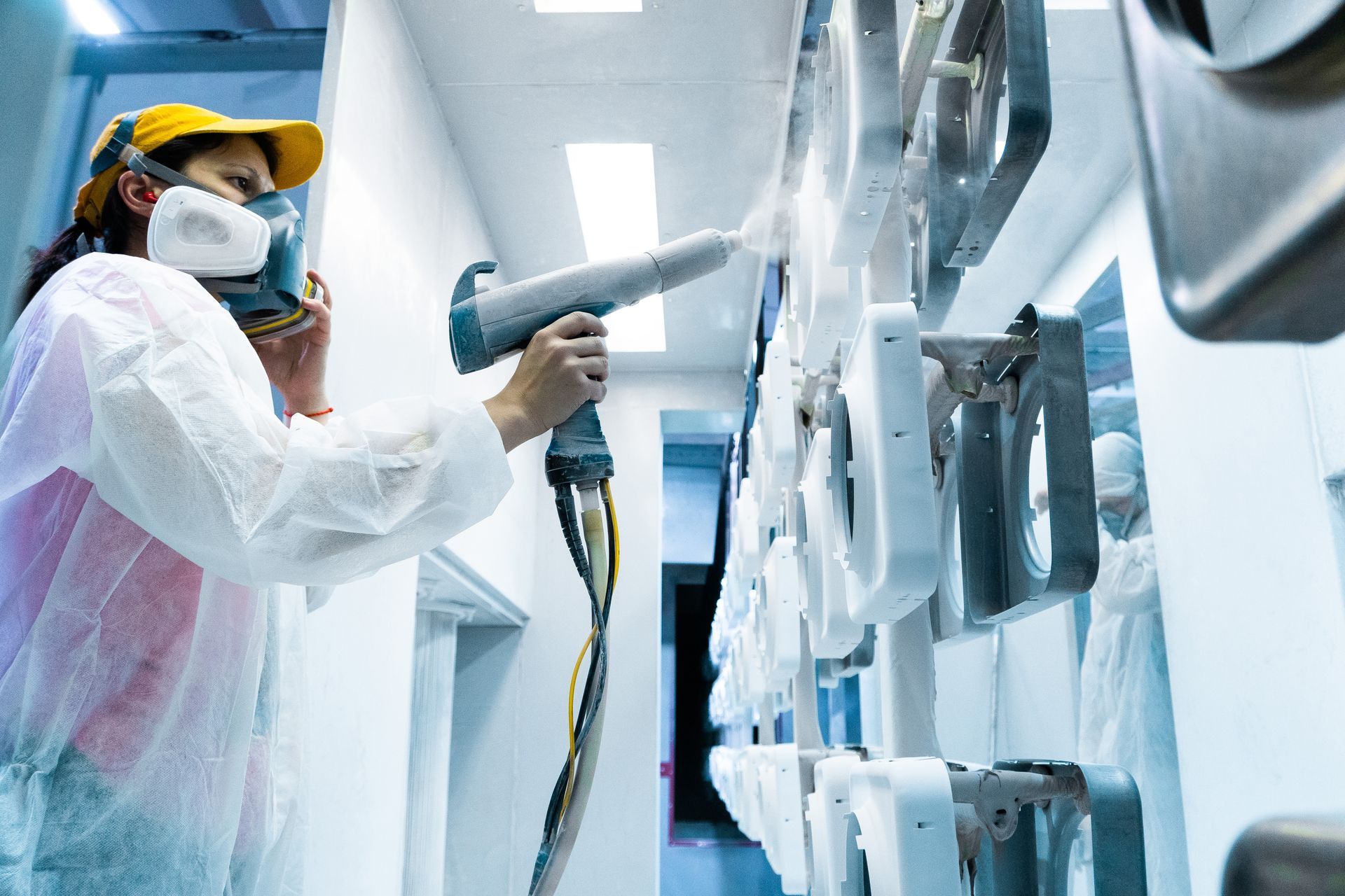Person in protective gear powder-coating metal parts in a white industrial setting.