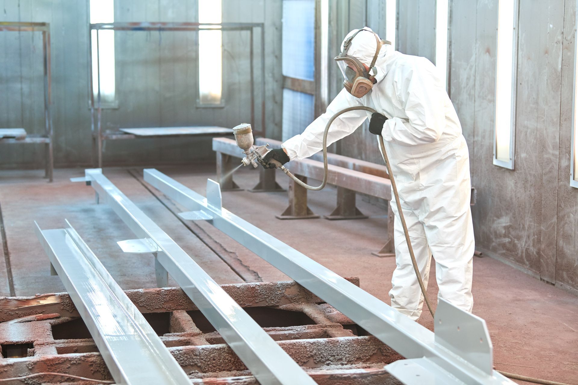 A worker in protective gear and a respirator spray-paints grey metal beams in a professional industrial workshop.